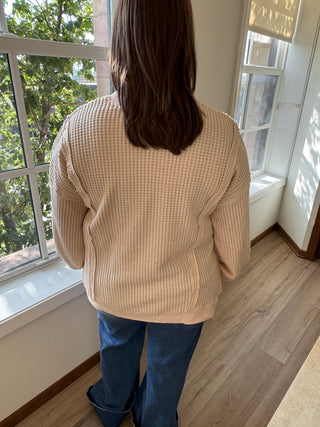 Person wearing a beige sweater and blue jeans standing indoors with a window in the background