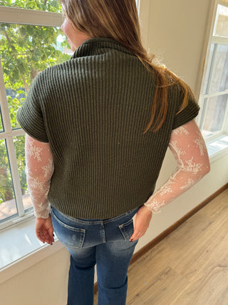Person wearing a green sweater and blue jeans indoors with a window in the background