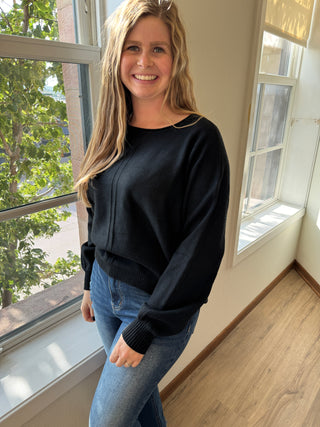 Person wearing a black sweater and blue jeans standing indoors with large windows.