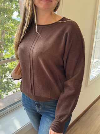Person wearing a brown sweater and blue jeans indoors with a window in the background