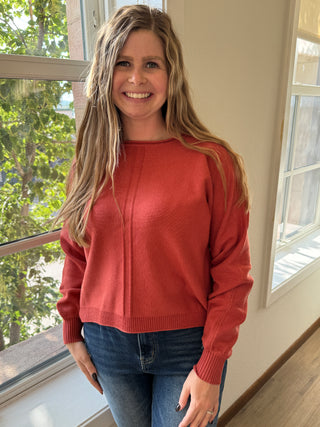 Woman wearing a red sweater and blue jeans indoors with a window in the background
