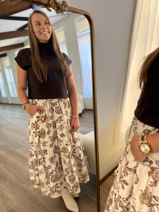 Woman standing in front of a mirror wearing a brown top and patterned skirt.