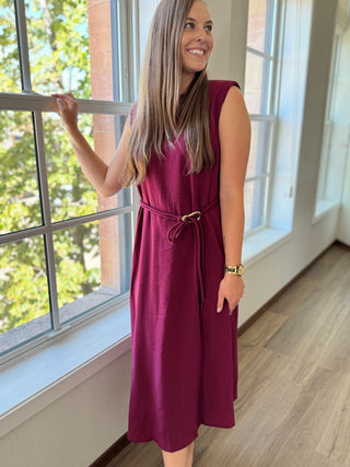 Woman in a maroon dress standing by a window with a view of greenery outside.