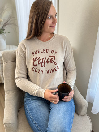 Lady sitting in chair holding coffee cup wearing a sweatshirt with words "Fueled by Coffee & Cozy Vibes".