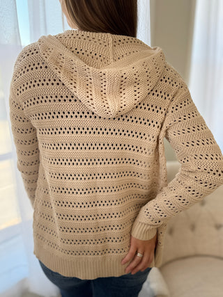 Back of Lady standing wearing a beige hooded cardigan.