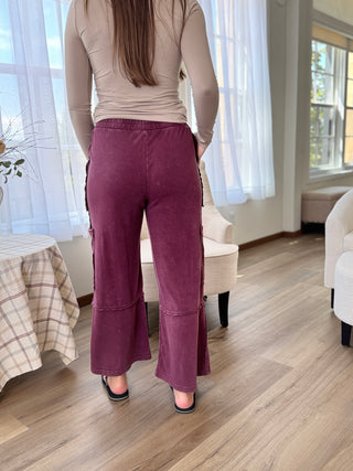 Person wearing purple pants and a beige top in a room with wooden flooring and white curtains.