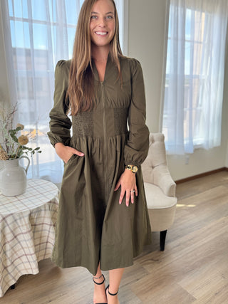 Woman wearing a green dress standing in a room with a table and chair.