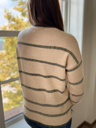Person wearing a beige sweater with black stripes, standing by a window with a view of trees.