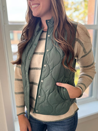 Woman wearing a green quilted vest over a beige sweater with a blurred indoor background
