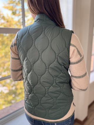 Person wearing a green quilted vest indoors with a blurred outdoor background