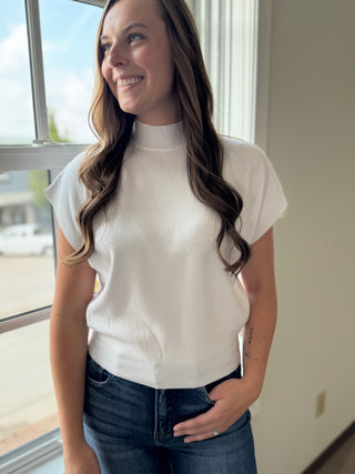 Woman wearing a white t-shirt and blue jeans standing by a window.
