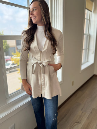 Woman wearing a white belted jacket and blue jeans standing by a window.