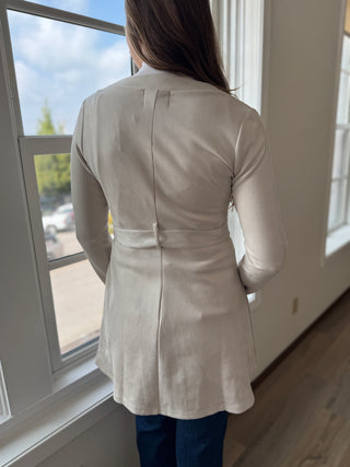 Person wearing a light-colored coat standing by a window with a view of a car outside.