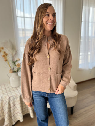 Woman wearing a beige cardigan and blue jeans in a room with a bed and window.