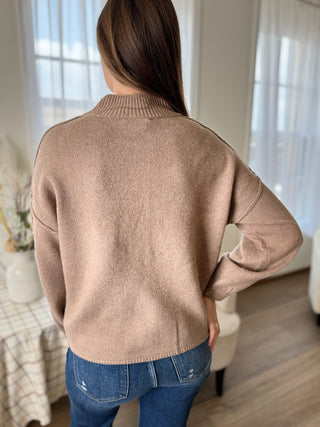 Person wearing a beige sweater and blue jeans in a room with light-colored walls and furniture.