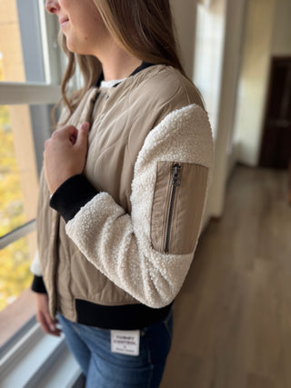 Person wearing a beige bomber jacket with fleece sleeves, standing indoors.