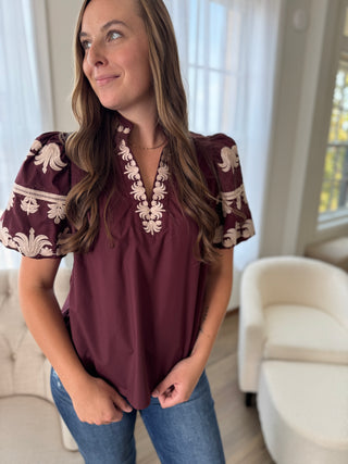 Woman wearing a burgundy blouse with floral embroidery in a living room.