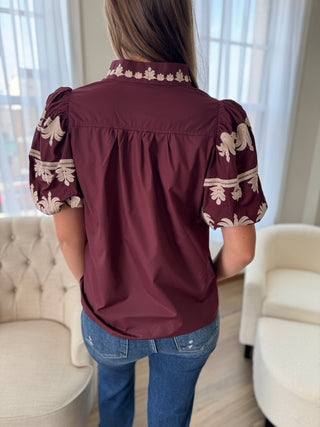 Person wearing a burgundy blouse with lace details in a room with white curtains and furniture.