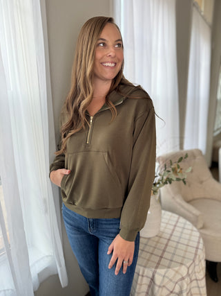 Woman wearing an olive green hoodie and blue jeans standing in a room with white curtains.