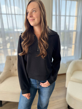 Woman wearing a black sweater and blue jeans standing in a room with large windows.