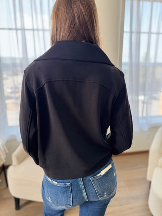 Person wearing a black jacket and blue jeans in a room with light-colored walls and furniture.