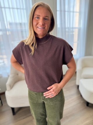 Woman wearing a brown turtleneck and green pants indoors.
