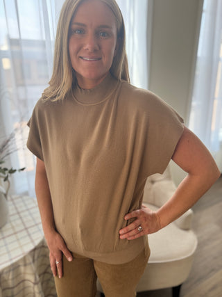 Woman wearing a beige turtleneck and pants in a bright room with large windows.