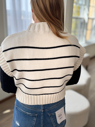 Person wearing a white sweater with black stripes and blue jeans indoors.