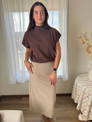 Woman wearing a brown top and beige skirt standing in a room with light curtains.