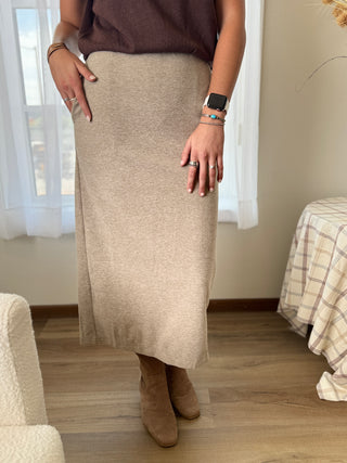 Person wearing a beige skirt and brown top in a room with a window and furniture.