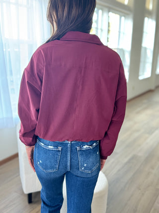 Person wearing a maroon jacket and blue jeans in a room with large windows.