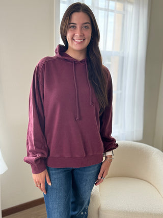 Woman wearing a maroon hoodie and blue jeans standing in a room with a window in the background.