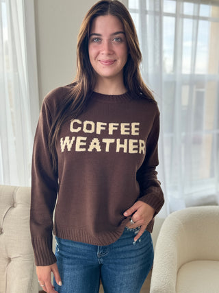 Woman wearing a brown sweater with 'COFFEE WEATHER' text in a home setting.