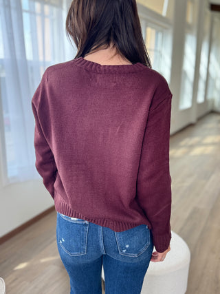 Person wearing a maroon sweater and blue jeans in a room with large windows.