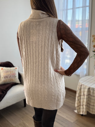 Person wearing a beige knitted vest in a room with furniture and decor.