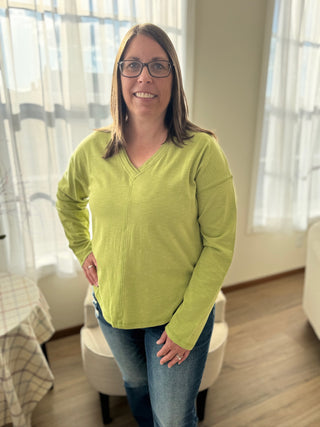 Woman wearing a green sweater standing in a room with light-colored curtains.