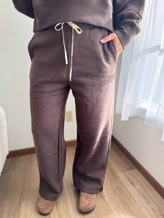 Person wearing brown sweatpants and a matching top in a room with light-colored walls and wooden floor.