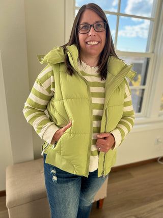 Woman wearing a green puffer vest over a striped shirt in a room with a window.
