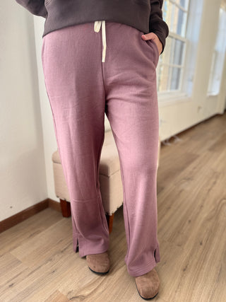 Person wearing mauve pants in a room with wooden flooring and a window.