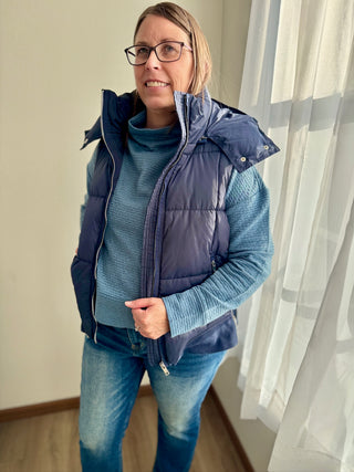 Person wearing a blue puffer vest over a blue sweater and jeans indoors.