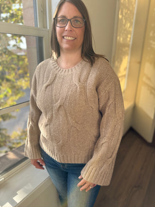 Woman wearing a beige cable knit sweater standing by a window with a view of trees.