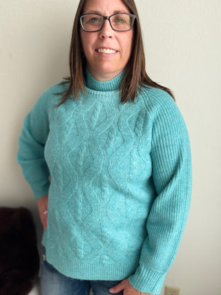 Person wearing a teal cable knit sweater against a plain background