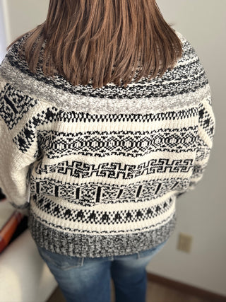 Person wearing a black and white patterned sweater with blue jeans indoors.