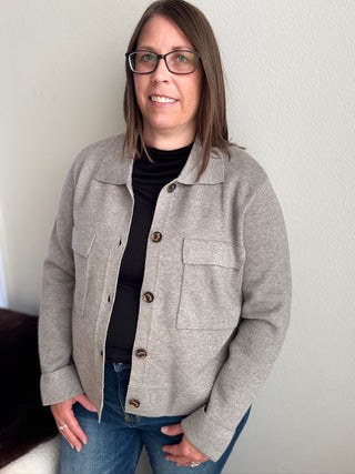 Person wearing a gray jacket over a black shirt and blue jeans against a plain background