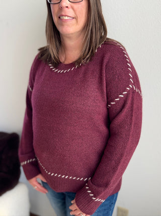 Person wearing a maroon sweater with white stitching details against a plain background