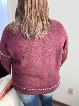 Person wearing a maroon sweater with white stitching details, standing indoors.