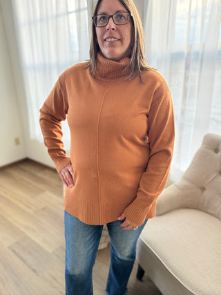 Person wearing a brown sweater and blue jeans standing in a room with light-colored curtains.