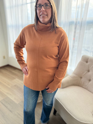 Woman wearing a brown sweater and blue jeans standing in a room with light-colored curtains.