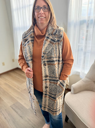 Woman wearing a patterned vest over a brown sweater in a room with curtains and a chair.
