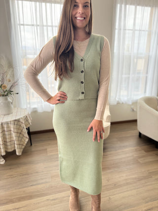Woman wearing a light green knitted dress with buttons in a room with white curtains and a table.